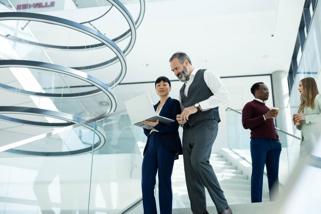 Business executive presenting laptop content to a colleague while walking down an upscale office staircase. Other colleagues